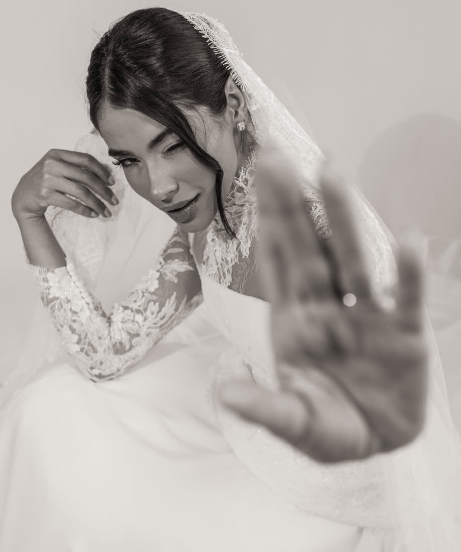 Noiva de véu e vestido de renda, em preto e branco, estendendo a mão para a câmera com o anel em primeiro plano